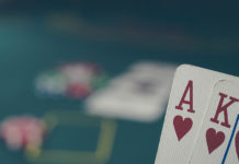 Podstawowe zasady pokera dla początkujących Zasady pokera - grafika promująca artykuł, przedstawiająca na pierwszym planie AK w kierach, a w tle stół pokerowy i żetony.