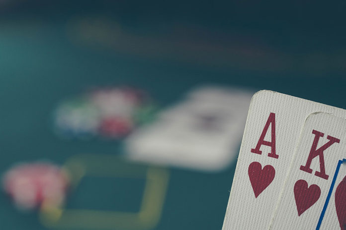Zasady pokera - grafika promująca artykuł, przedstawiająca na pierwszym planie AK w kierach, a w tle stół pokerowy i żetony.